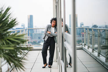 Porträt einer schwarzen Geschäftsfrau, die mit verschränkten Armen auf einer Terrasse vor dem Panorama einer Londoner Dachterrasse steht; Kopierraum. - HPIF30232