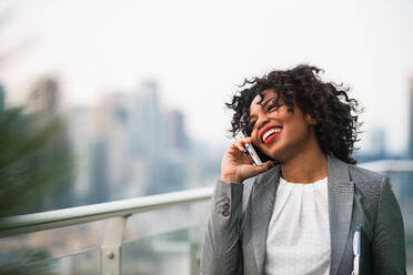 A portrait of black businesswoman with smartphone standing on a terrace against London rooftop view panorama, making a phone call. Copy space. - HPIF30239