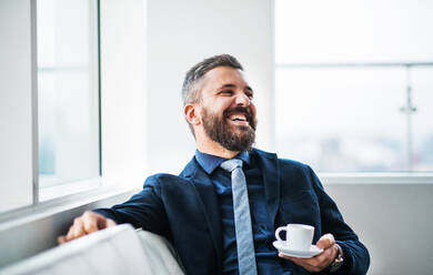 Porträt eines Geschäftsmannes, der mit einer Tasse Kaffee auf einem weißen Sofa in einem modernen Büro sitzt und lacht. Kopierraum. - HPIF30240