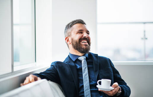 Porträt eines Geschäftsmannes, der mit einer Tasse Kaffee auf einem weißen Sofa in einem modernen Büro sitzt und lacht. Kopierraum. - HPIF30240