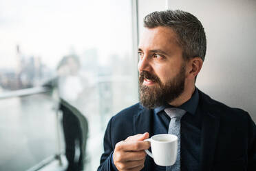 Nahaufnahme eines Geschäftsmannes mit einer Tasse Kaffee, der aus einem Fenster in einem Büro schaut. - HPIF30242