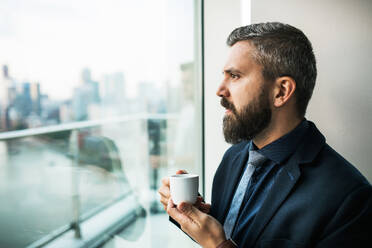 Nahaufnahme eines Geschäftsmannes mit einer Tasse Kaffee, der aus einem Fenster in einem Büro schaut. - HPIF30243