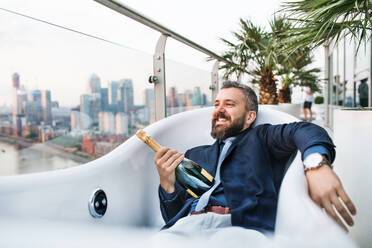 Geschäftsmann mit einer Flasche Champagner in einem leeren Whirlpool liegend, im Hintergrund das Panorama von London. - HPIF30245