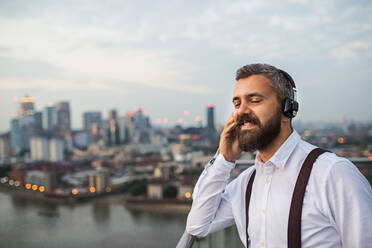 A businessman with headphones standing against London view panorama, listening to music. Copy space. - HPIF30269