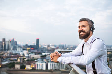 Ein Geschäftsmann mit Kopfhörern steht vor dem Panorama Londons und hört Musik. Raum kopieren. - HPIF30270