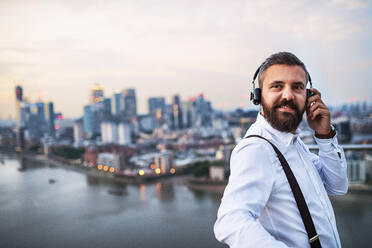 Ein Geschäftsmann mit Kopfhörern steht vor dem Panorama Londons und hört Musik. Raum kopieren. - HPIF30271