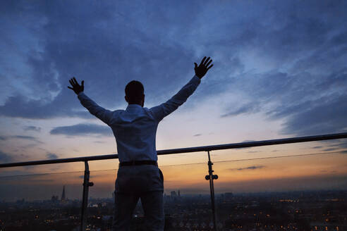 Rückansicht eines Hipster-Geschäftsmannes, der in der Abenddämmerung vor dem Panorama Londons steht und die Hände hebt. Raum kopieren. - HPIF30286