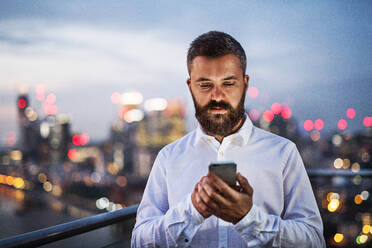 A portrait of businessman with smartphone standing against night London rooftop view panorama at sunset, text messaging. Copy space. - HPIF30288