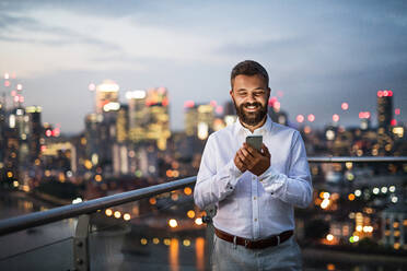 A portrait of businessman with smartphone standing against night London rooftop view panorama. Copy space. - HPIF30289