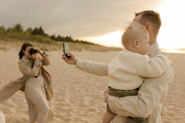 Ein Vater hält einen Moment mit seinem Sohn fest, während eine Frau denselben Moment durch ihr Kameraobjektiv am Strand festhält - VIVF00950