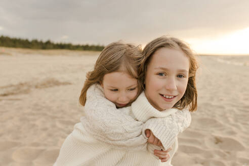 Glückliches Mädchen, das seine Schwester am Strand huckepack nimmt - VIVF00983