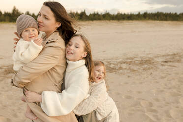 Smiling daughters hugging mother at beach - VIVF00989