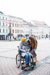 A teenager girl with disabled grandmother in wheelchair outdoors on the street in winter, talking. - HPIF30345