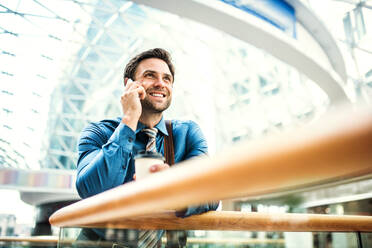 Ein junger Geschäftsmann mit Kaffee und Smartphone steht in einem modernen Gebäude und führt ein Telefongespräch. - HPIF30355