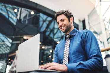 Ein junger, gut aussehender Geschäftsmann sitzt auf einer Bank in einem modernen Gebäude und benutzt einen Laptop. - HPIF30367