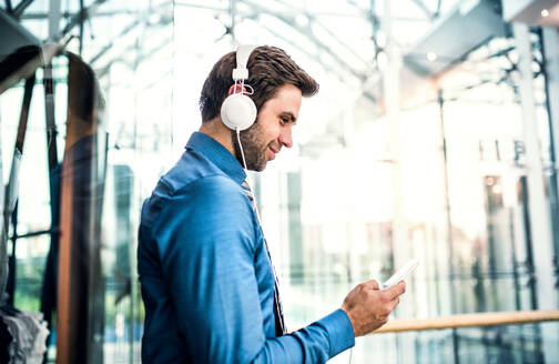 Ein junger Geschäftsmann mit Smartphone und Kopfhörern steht in einem modernen Einkaufszentrum und hört Musik. - HPIF30370