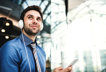 A young businessman with smartphone and headphones standing in a modern shopping center, listening to music. Copy space. - HPIF30372