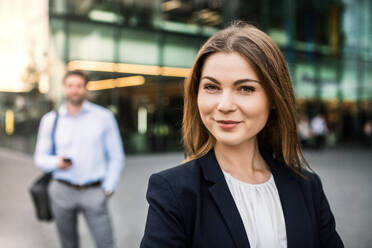 A portrait of a young businesswoman standing outdoors in front of a building, taking selfie. A man walking in the background, using smartphone. - HPIF30387