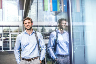 A young businessman with a bag walking in front of a modern building, hands in pockets. - HPIF30394