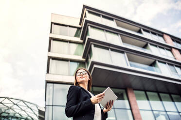 A low angle view of young businesswoman using tablet outdoors in front of a building in a city. Copy space. - HPIF30395