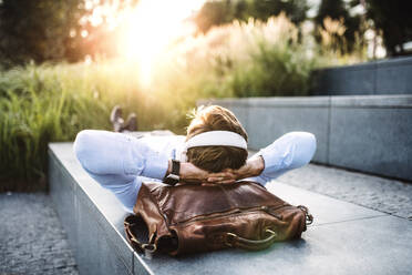 A businessman with headphones, lying on the steps outdoors at sunset, a head resting on a laptop bag. - HPIF30403