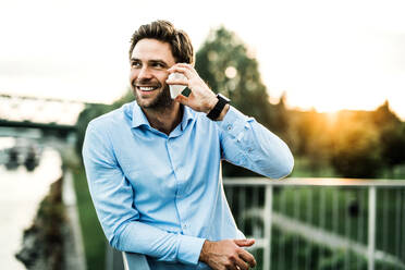 Ein junger Geschäftsmann mit Smartphone steht bei Sonnenuntergang auf einer Brücke, stützt sich auf ein Geländer und telefoniert. - HPIF30416