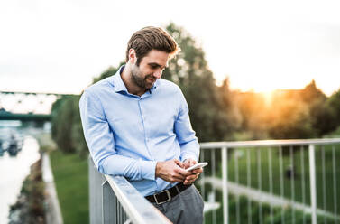 A young businessman with smartphone standing on a bridge at sunset, leaning on a railing. Copy space. - HPIF30417