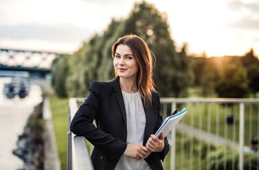A young businesswoman standing on the river promenade outdoors at sunset. - HPIF30420