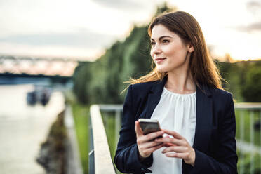 A young businesswoman standing outdoors on the river bank, using smartphone. - HPIF30424