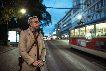 Mature businessman with suitcase waiting for a tram or a trolley car in the evening in Prague city. - HPIF30447