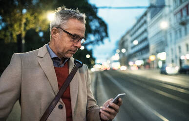 Ein reifer Geschäftsmann mit Smartphone steht abends auf einer Straße und schreibt eine SMS. Platz kopieren. - HPIF30448
