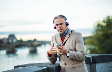 Mature handsome businessman with headphones and smartphone standing by river Vltava in Prague city, listening to music and singing. Copy space. - HPIF30465