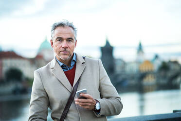 Mature handsome businessman with smartphone standing by river Vltava in Prague city. Copy space. - HPIF30466