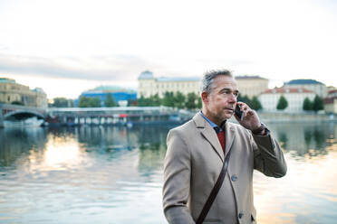 Älterer, gut aussehender Geschäftsmann mit Smartphone, der am Fluss Moldau in Prag steht und telefoniert. Kopierraum. - HPIF30471