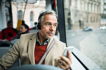 Reifer, müder Geschäftsmann mit Smartphone und Heaphones, der mit dem Bus in der Stadt unterwegs ist und Musik hört. - HPIF30480