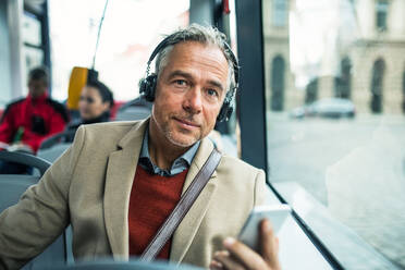 Reifer, müder Geschäftsmann mit Smartphone und Heaphones, der mit dem Bus in der Stadt unterwegs ist und Musik hört. - HPIF30481
