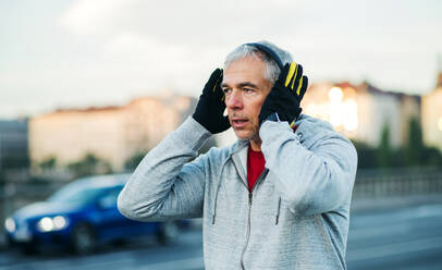 Älterer männlicher Läufer mit Handschuhen, der im Freien auf einer Brücke in Prag Kopfhörer aufsetzt. - HPIF30495