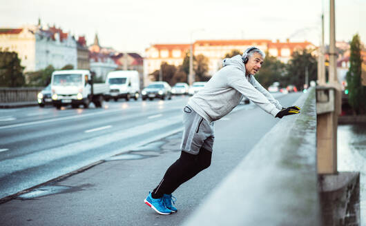 Ein fitter reifer männlicher Läufer mit Kopfhörern, der sich im Freien auf einer Brücke in Prag dehnt und Musik hört. - HPIF30499