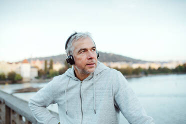 Reifer männlicher Läufer mit schwarzen Kopfhörern im Freien auf der Brücke in der Stadt Prag, Musik hörend. - HPIF30501