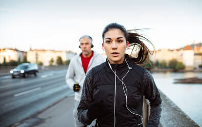 A fit sporty couple with headphones running outdoors on the bridge in Prague city, listening to music. - HPIF30508