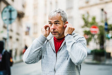 A portrait of an active mature man standing outdoors in city, putting earphones in his ears. - HPIF30515