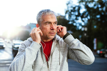 A portrait of an active mature man standing outdoors in city, putting earphones in his ears. - HPIF30524