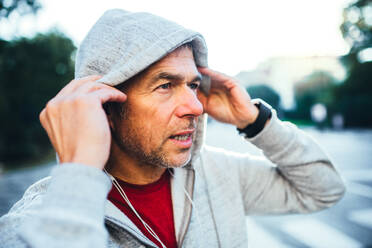 Ein Porträt eines aktiven, fröhlichen, reifen Mannes mit Kopfhörern, der draußen in der Stadt steht und sich eine graue Kapuze über den Kopf zieht. - HPIF30531