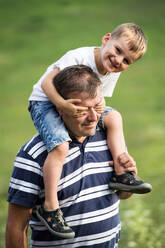 Ein junger Vater, der seinen kleinen Sohn an einem Sommertag in der Natur huckepack nimmt, wobei der Junge die Augen des Mannes verdeckt. - HPIF30534
