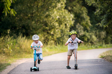 Zwei fröhliche kleine Jungen mit Helm fahren an einem Sommertag Roller auf einer Straße im Park. - HPIF30540