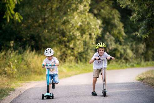 Two cheerful small boys with a helmet riding scooters on a road in park on a summer day. - HPIF30540