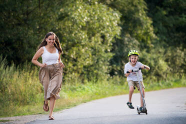 Ein fröhlicher kleiner Junge, der Roller fährt und seine Mutter, die an einem Sommertag auf einer Straße im Park läuft. - HPIF30541