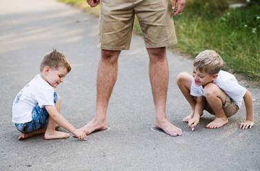 Two small sons playing with unrecognizable father on a road in park on a summer day, drawing with chalk. - HPIF30545