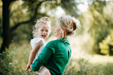 Schöne junge Mutter in grüner sonniger Sommernatur, die ihre süße kleine Tochter in den Armen hält und Spaß hat. - HPIF30563