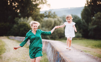 Young mother in nature with small daughter, having fun. A girl running on a concrete wall, holding hands. - HPIF30591